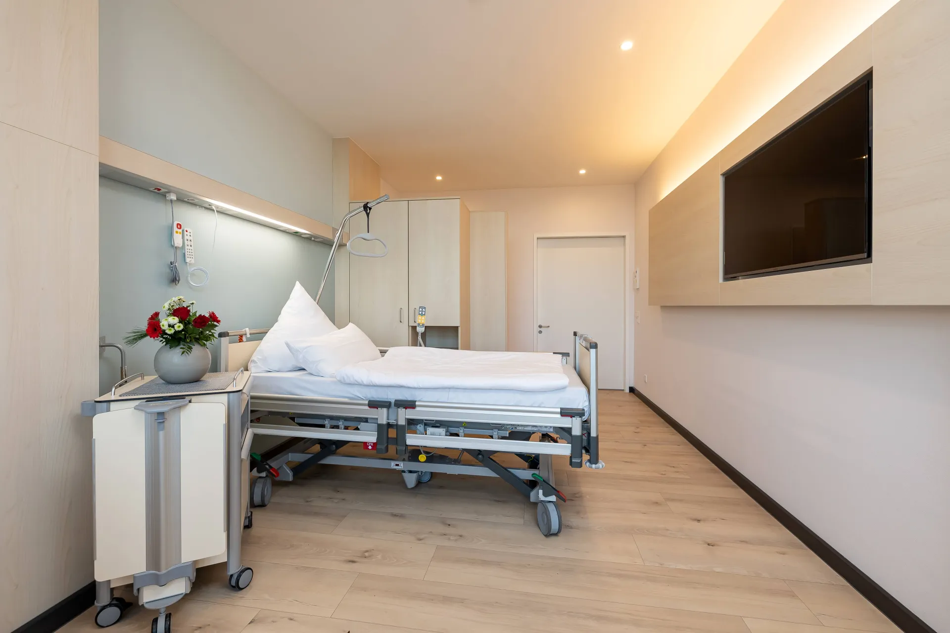 Hospital room with a bed, bedside table with a vase and flowers, a large wall-mounted television and a wooden floor